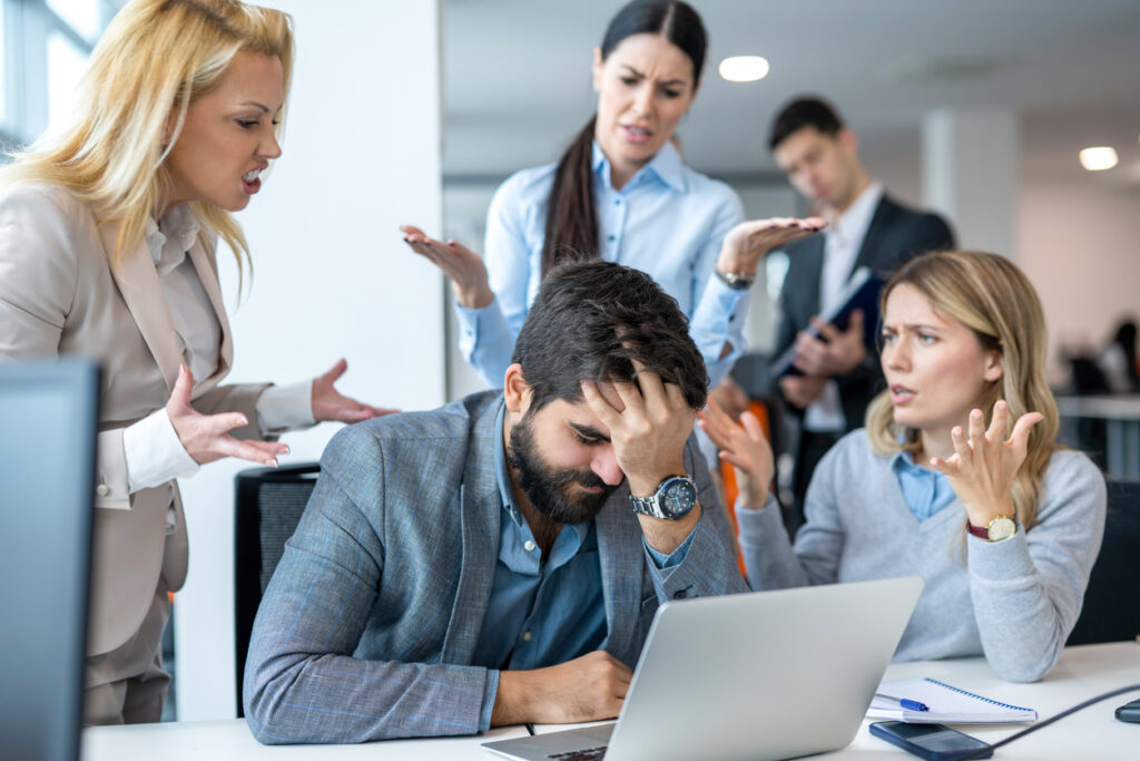 Frustrated businessman covering face with hand, having serious problems while his co-workers making him feels guilty for not doing his job properly at office. Business conflict and overwork concept.