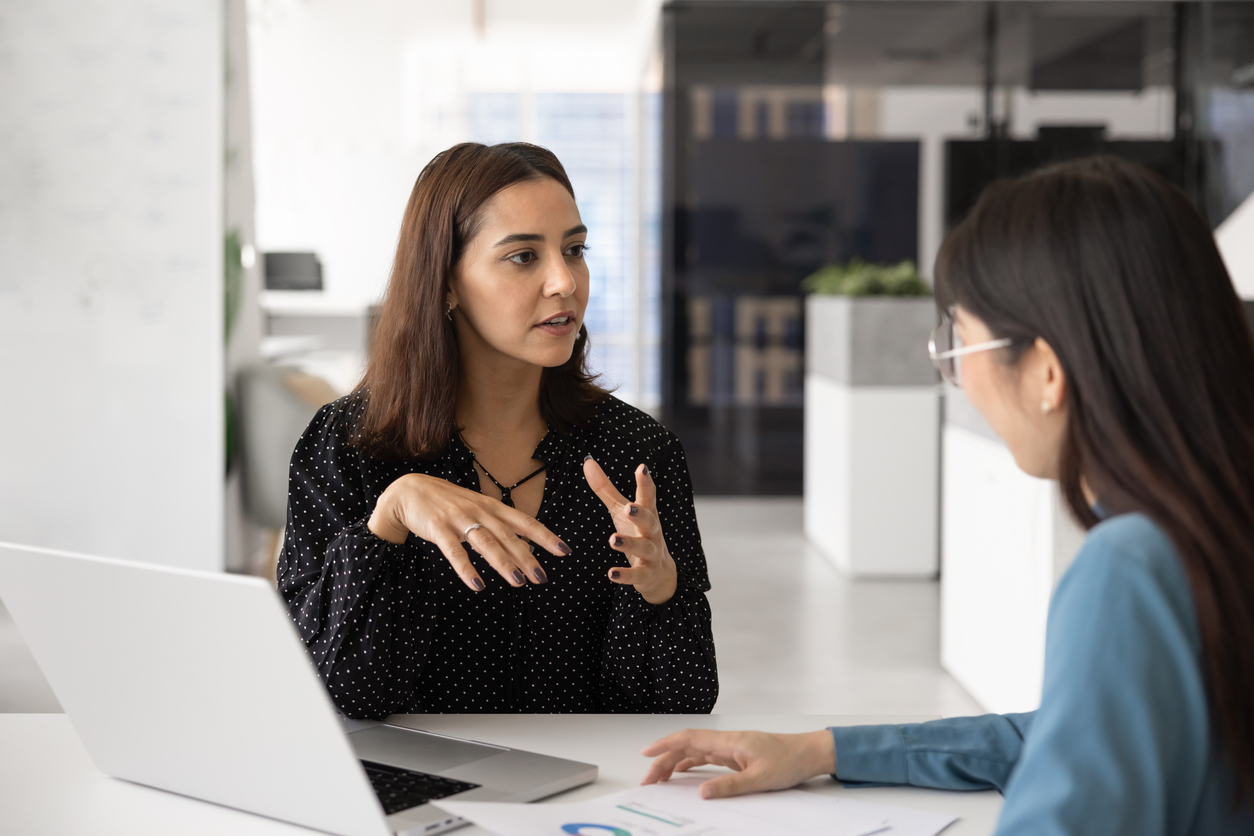 Two coworkers having a conversation at a desk with a laptop.